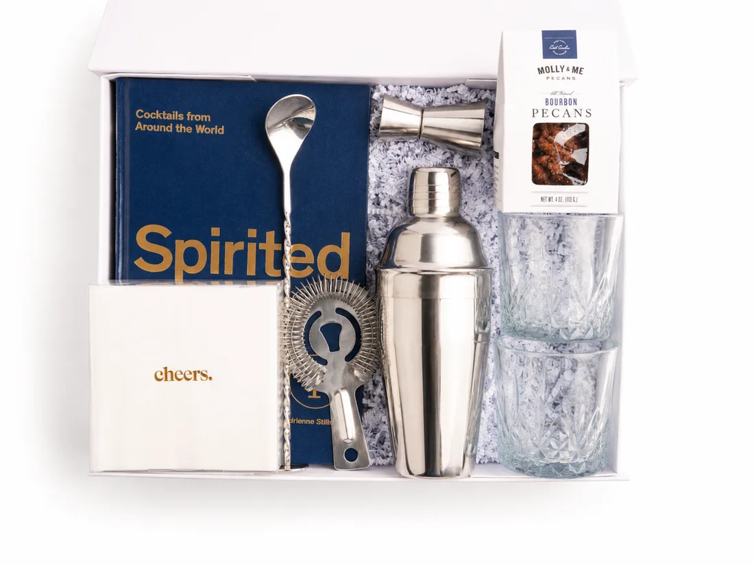 White gift box with a cocktail shaker, jigger, strainer, cut-glass tumblers, bourbon pecans and a cocktail book