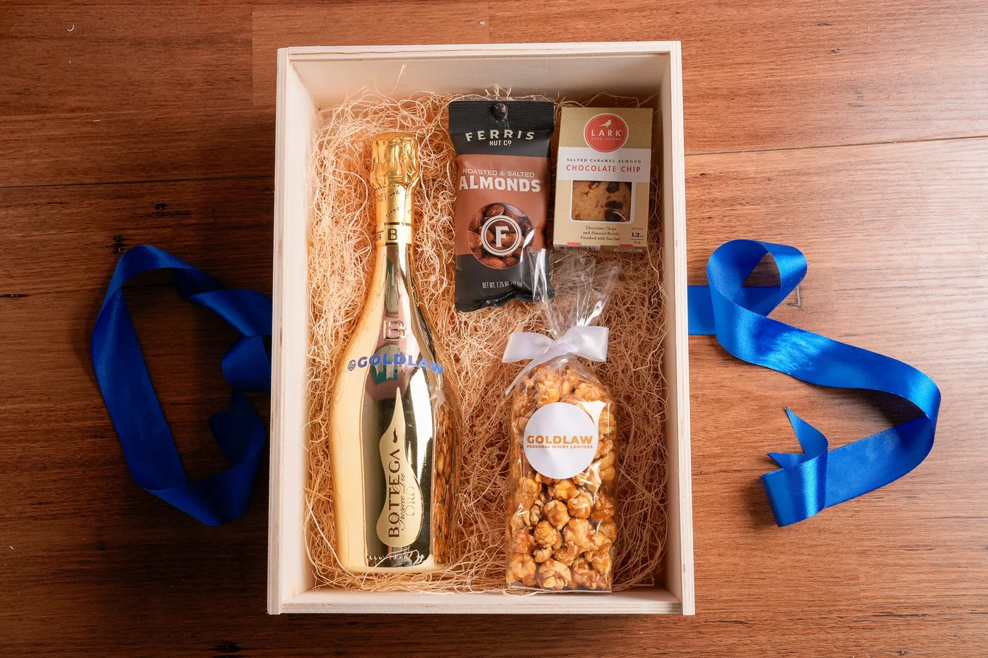 Timber crate lined with wood wool holding a gold prosecco bottle, almonds, cookies and caramel popcorn, branded for a law firm and finished with blue ribbon