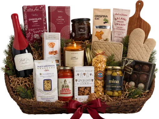 Wicker basket with a bottle of red wine, chocolates, salami, olive crackers, fig spread, pasta, a wooden cutting board, oven mitt and a lit candle, finished with a red bow