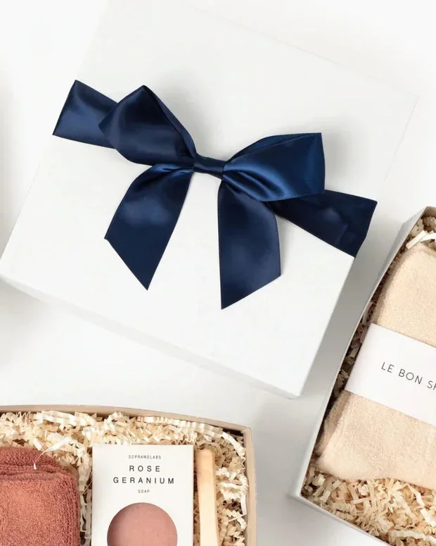 A white gift box finished with a wide navy satin bow, resting beside two open boxes packed with towels and soap
