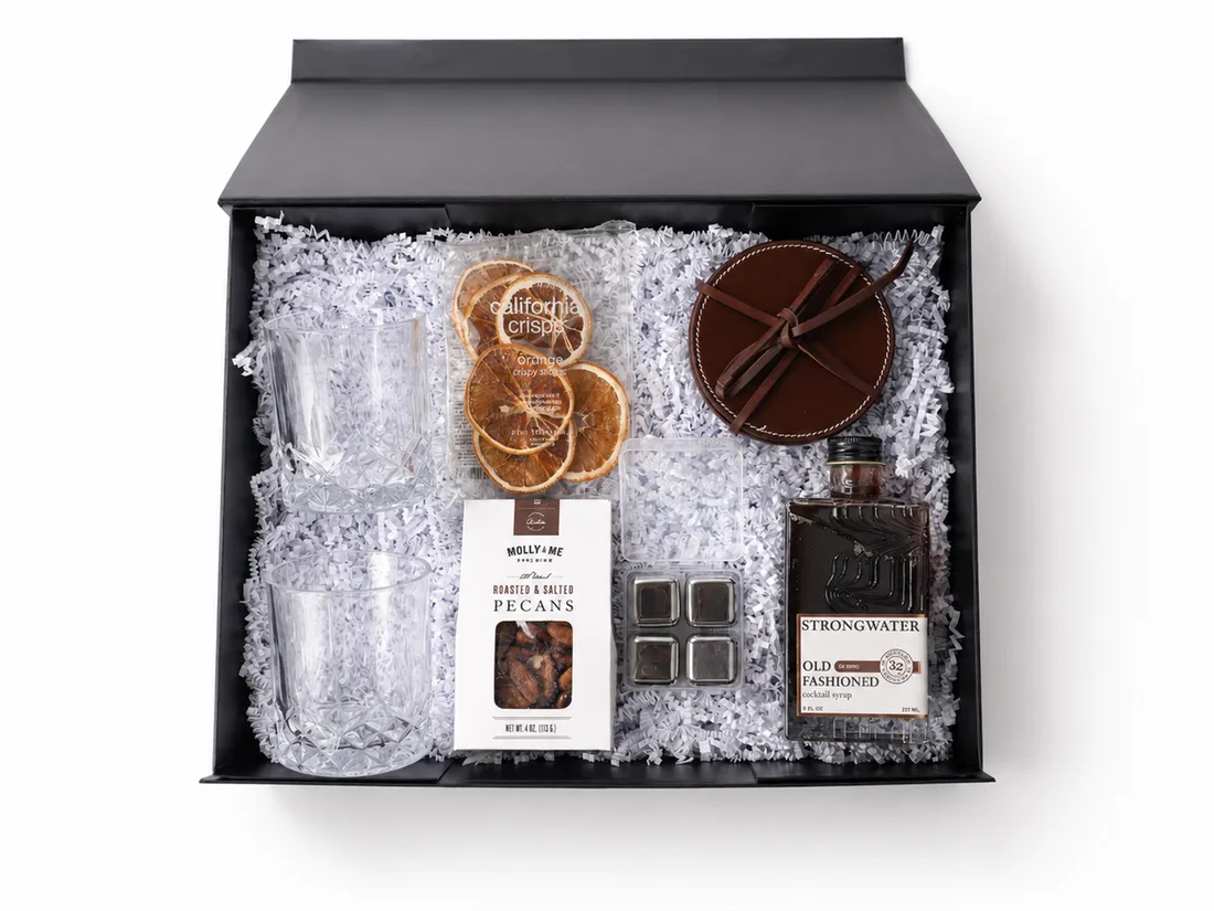 Black gift box holding cut-glass tumblers, whiskey stones, old-fashioned cocktail syrup, roasted pecans, dried orange slices and a leather coaster set